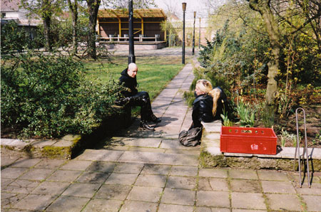 Zwei schwarz gekleidete Gestalten sitzen am Wegesrand eines Parks.